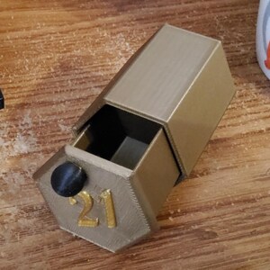 May include: A gold-colored, hexagonal box with a drawer pulled open. The number "21" is printed in gold on the front of the box. A black knob is on the drawer. The box is sitting on a wooden surface.