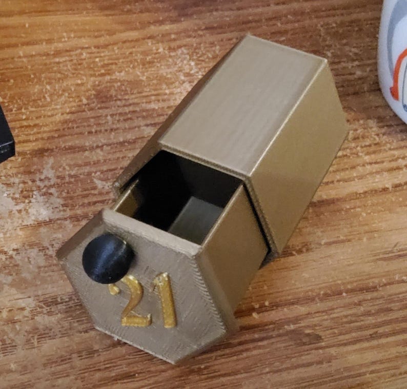 May include: A gold-colored, hexagonal box with a drawer pulled open. The number "21" is printed in gold on the front of the box. A black knob is on the drawer. The box is sitting on a wooden surface.