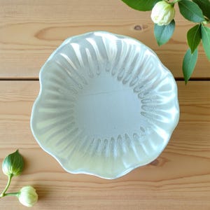 May include: A small, off-white, scalloped-edge bowl with a textured, ridged interior. The bowl is set on a light brown wooden surface, with a sprig of green leaves and white buds in the background.