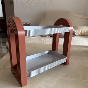 May include: A two-tiered decorative shelf with a terracotta-colored frame and two gray shelves. The shelves have a ribbed texture. The frame has an arched design on the sides. The shelf is designed to hold small items.