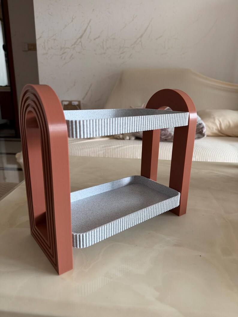 May include: A two-tiered decorative shelf with a terracotta-colored frame and two gray shelves. The shelves have a ribbed texture. The frame has an arched design on the sides. The shelf is designed to hold small items.