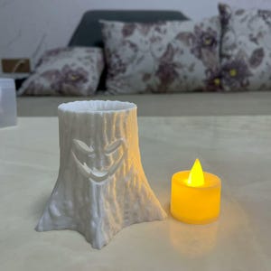 Puede incluir: Un portavelas blanco con forma de toc&oacute;n de &aacute;rbol con una cara sonriente tallada. Una peque&ntilde;a vela amarilla que funciona con pilas se encuentra junto al soporte, emitiendo un brillo c&aacute;lido. El portavelas es un art&iacute;culo decorativo.