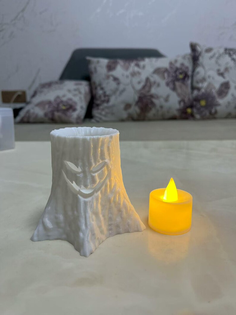 Puede incluir: Un portavelas blanco con forma de toc&oacute;n de &aacute;rbol con una cara sonriente tallada. Una peque&ntilde;a vela amarilla que funciona con pilas se encuentra junto al soporte, emitiendo un brillo c&aacute;lido. El portavelas es un art&iacute;culo decorativo.