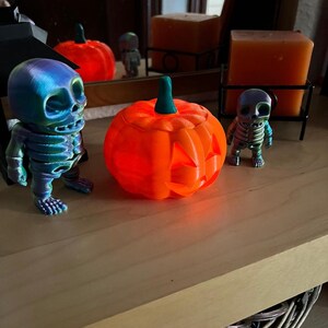 May include: An orange, 3D-printed jack-o'-lantern with a green stem sits on a wooden surface, flanked by two skeleton figurines. The skeletons have a rainbow-colored finish. A mirror in the background reflects the scene, including two orange candles.