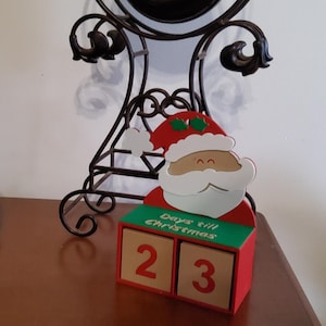 May include: A wooden Christmas countdown calendar shaped like Santa Claus. The red and green calendar displays "Days till Christmas" and shows the number 23. The Santa figure has a white beard and a red hat with holly.