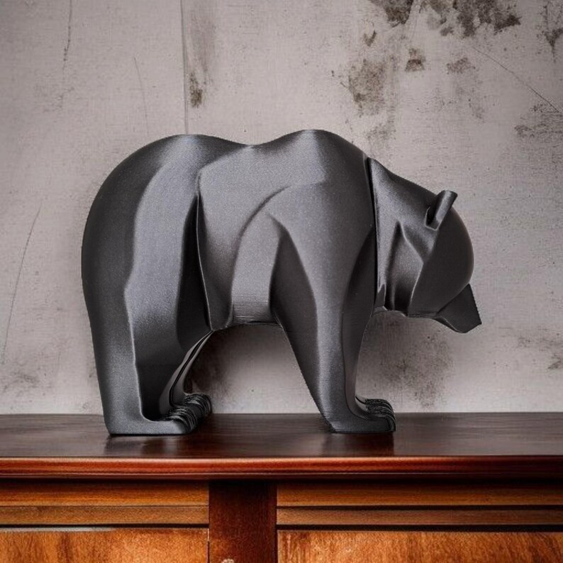 May include: A dark gray bear sculpture with a geometric design. The bear is standing on all fours, with its head slightly lowered. The sculpture is placed on a wooden surface against a textured gray wall.
