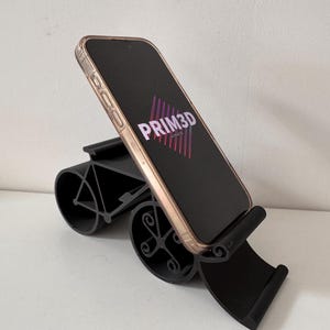 May include: A black phone stand with a curved design, holding a smartphone. The phone has a gold-colored frame and displays the text "PRIM3D" in pink and purple. The stand is on a white surface.