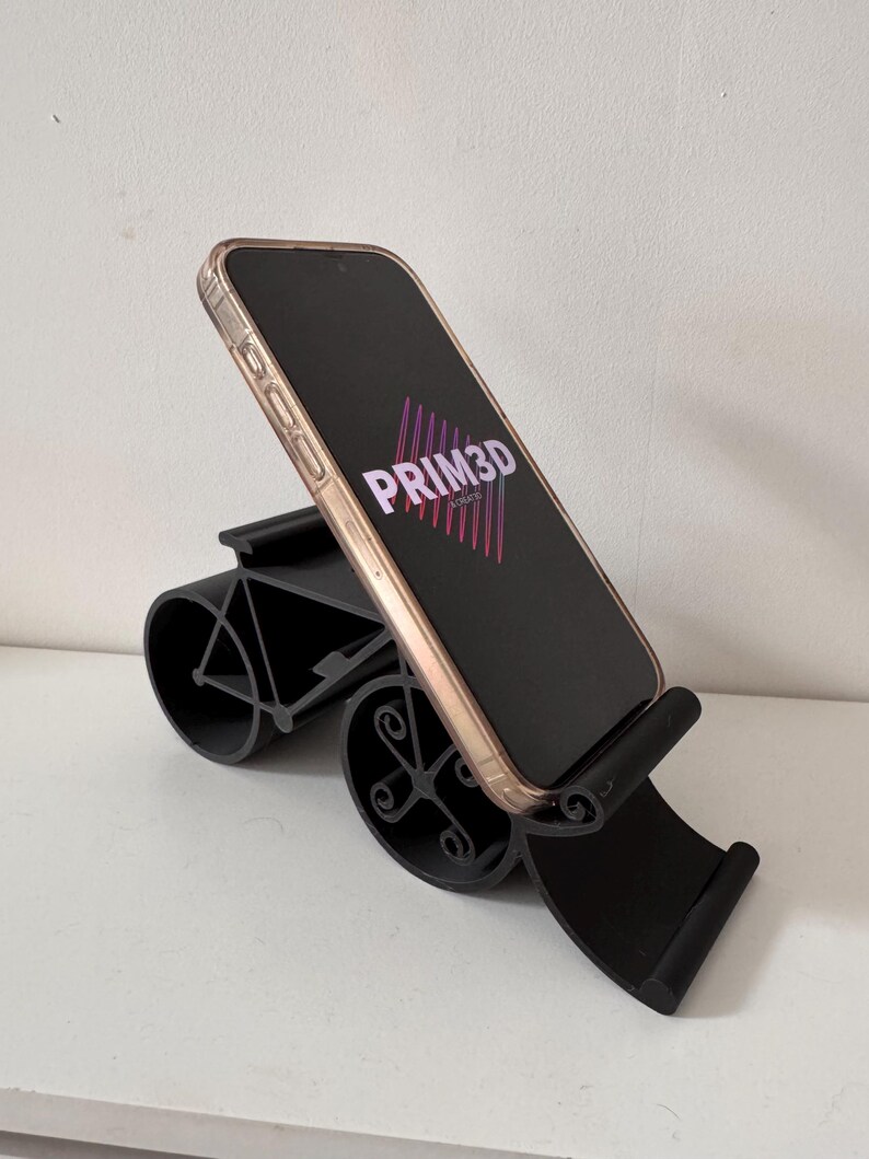 May include: A black phone stand with a curved design, holding a smartphone. The phone has a gold-colored frame and displays the text "PRIM3D" in pink and purple. The stand is on a white surface.