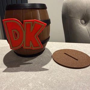 May include: A brown barrel-shaped piggy bank with black bands and a red and gold "DK" logo. A brown, oval-shaped lid with a slot sits next to the bank. The bank is on a light gray surface.