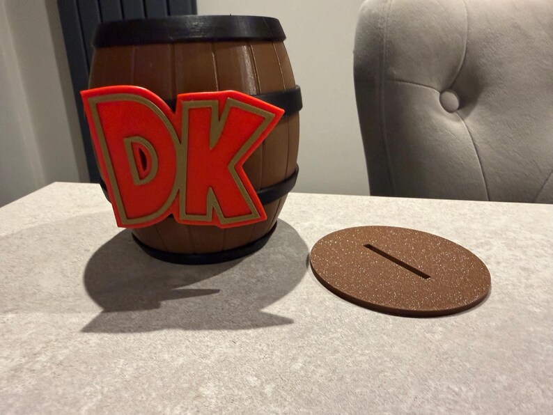 May include: A brown barrel-shaped piggy bank with black bands and a red and gold "DK" logo. A brown, oval-shaped lid with a slot sits next to the bank. The bank is on a light gray surface.