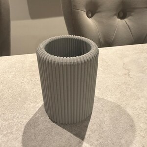 May include: A cylindrical, light gray container with vertical ribbed detailing. The container is made of a solid material and casts a shadow on the surface it rests on. The background includes a chair and a table.