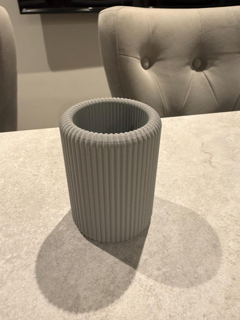 May include: A cylindrical, light gray container with vertical ribbed detailing. The container is made of a solid material and casts a shadow on the surface it rests on. The background includes a chair and a table.