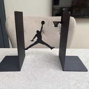 May include: Black metal bookends featuring a silhouette of a basketball player mid-air, about to dunk the ball. The bookends have a rectangular base and a vertical support. The basketball hoop is also included in the design.
