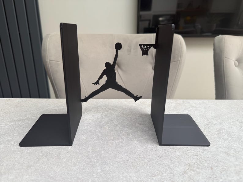 May include: Black metal bookends featuring a silhouette of a basketball player mid-air, about to dunk the ball. The bookends have a rectangular base and a vertical support. The basketball hoop is also included in the design.