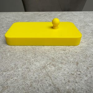 K&ouml;nnte beinhalten: Ein leuchtend gelbes rechteckiges Objekt mit einem kleinen, abgerundeten Knopf oben. Das Objekt scheint ein Deckel oder ein Beh&auml;lter zu sein, m&ouml;glicherweise f&uuml;r die Aufbewahrung oder zu dekorativen Zwecken. Die Farbe ist durchgehend.