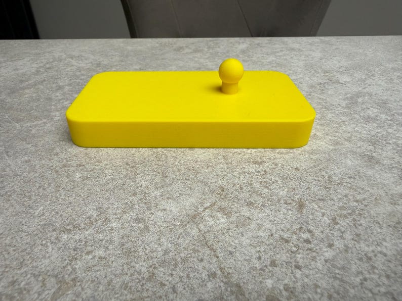K&ouml;nnte beinhalten: Ein leuchtend gelbes rechteckiges Objekt mit einem kleinen, abgerundeten Knopf oben. Das Objekt scheint ein Deckel oder ein Beh&auml;lter zu sein, m&ouml;glicherweise f&uuml;r die Aufbewahrung oder zu dekorativen Zwecken. Die Farbe ist durchgehend.