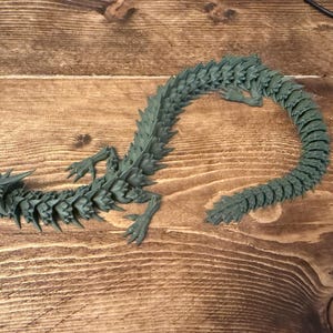 May include: A dark teal articulated dragon figurine. The dragon has a detailed, segmented body, legs, and a curved tail. The figurine is lying on a wooden surface.