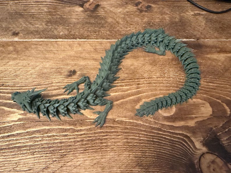 May include: A dark teal articulated dragon figurine. The dragon has a detailed, segmented body, legs, and a curved tail. The figurine is lying on a wooden surface.