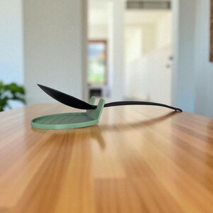 May include: A sage green spoon rest with a black spoon. The spoon rest has a circular base with a raised edge and a small handle rest. The spoon is black and has a long, thin handle. The spoon rest is on a wooden table.
