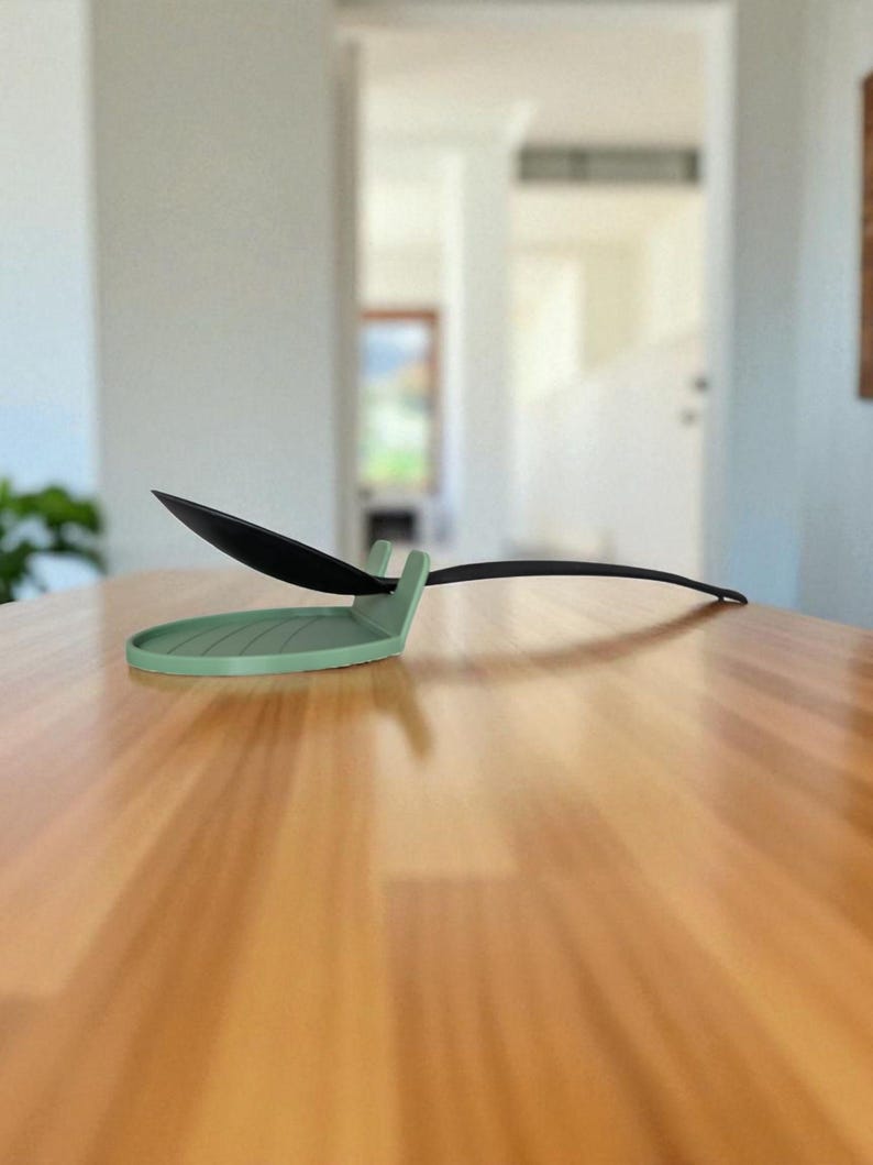 May include: A sage green spoon rest with a black spoon. The spoon rest has a circular base with a raised edge and a small handle rest. The spoon is black and has a long, thin handle. The spoon rest is on a wooden table.