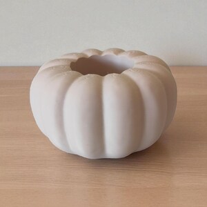 May include: A small, off-white ceramic pumpkin-shaped vase. The vase has a wide opening and a ribbed exterior, resembling the shape of a pumpkin. The vase is sitting on a light-colored wooden surface.
