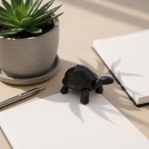 May include: A black geometric wooden turtle figurine sits on a white notepad. A succulent plant in a gray pot and a silver pen are also visible. The turtle has a polygonal design.