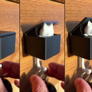 May include: A black, square key holder with a hinged lid, featuring a white cat figurine that pops up when the lid is opened. The key holder is mounted on a white hook, with a hand reaching for a set of keys.
