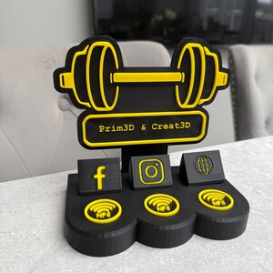 May include: Black and yellow dumbbell-shaped business card holder with the text "Prim3D & Creat3D". Features social media icons for Facebook, Instagram, and a globe, along with NFC symbols. The design is 3D printed.