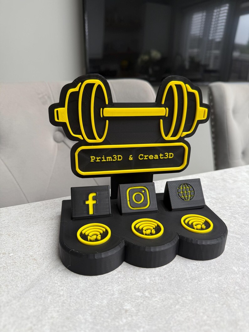 May include: Black and yellow dumbbell-shaped business card holder with the text "Prim3D & Creat3D". Features social media icons for Facebook, Instagram, and a globe, along with NFC symbols. The design is 3D printed.