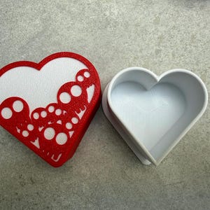 May include: A red and white heart-shaped box with a decorative cutout design. The box is open, revealing a white interior. The box is made of plastic and sits on a gray surface. The design is a series of circles.