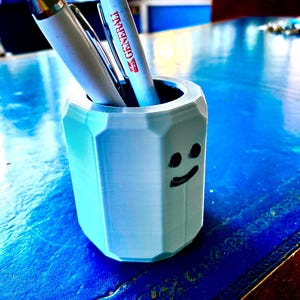 May include: A light blue, 3D-printed pen holder with a smiling face design. Two pens are inside the holder, one with a red tip and the other with the word "Generali" printed on it. The holder sits on a blue surface.