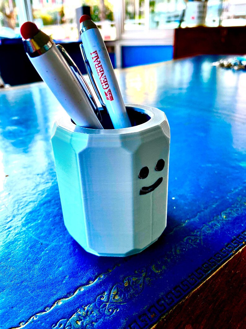 May include: A light blue, 3D-printed pen holder with a smiling face design. Two pens are inside the holder, one with a red tip and the other with the word "Generali" printed on it. The holder sits on a blue surface.