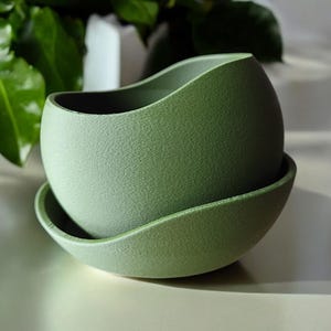 May include: A sage green ceramic planter with a textured finish, sitting on a matching saucer. The rounded shape of the planter contrasts with the curved saucer. The background features green leaves and a white surface.