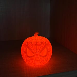 May include: Glowing orange pumpkin-shaped Halloween decoration. The pumpkin features a Spider-Man mask design with a web pattern and eye details. The decoration is lit from within, creating a warm, inviting glow.