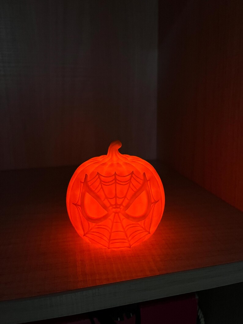 May include: Glowing orange pumpkin-shaped Halloween decoration. The pumpkin features a Spider-Man mask design with a web pattern and eye details. The decoration is lit from within, creating a warm, inviting glow.