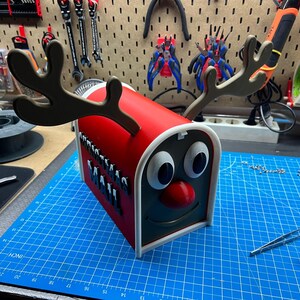 May include: A red and white decorative mailbox shaped like a reindeer with brown antlers, large white eyes, and a red nose. The mailbox has a black face and a smiling mouth. The mailbox is sitting on a blue cutting mat.