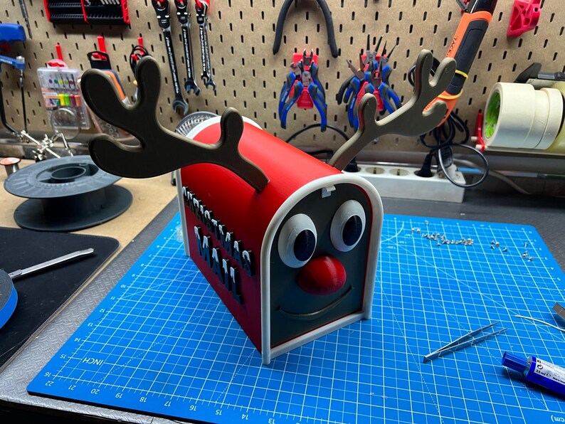 May include: A red and white decorative mailbox shaped like a reindeer with brown antlers, large white eyes, and a red nose. The mailbox has a black face and a smiling mouth. The mailbox is sitting on a blue cutting mat.