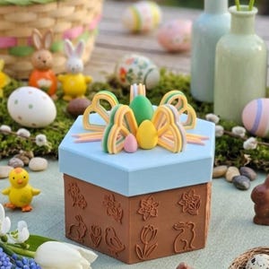 May include: A light blue and brown Easter-themed gift box with embossed floral and bunny designs. The box is topped with decorative eggs and a bow. Easter eggs, bunnies, and flowers surround the box, creating a festive scene.