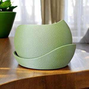 May include: A light green, textured ceramic plant pot with a matching saucer. The pot has a unique, curved design and sits on a wooden surface. Another green pot is visible in the background. Suitable for indoor plants.