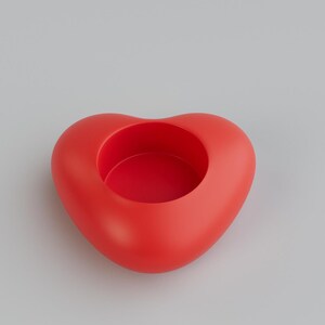 May include: A red heart-shaped container with a circular opening in the center. The container is a solid, smooth, and glossy red. The background is a plain, light gray.