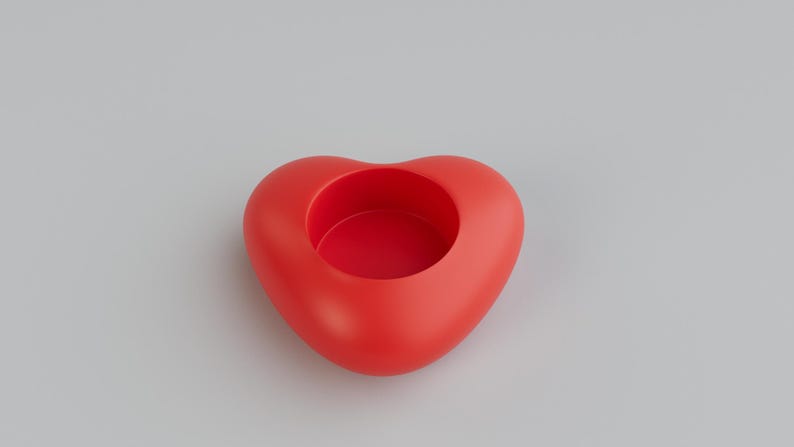 May include: A red heart-shaped container with a circular opening in the center. The container is a solid, smooth, and glossy red. The background is a plain, light gray.