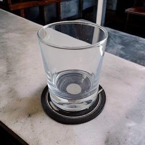 May include: A clear, empty drinking glass sits on a black coaster with a white circular design. The glass is partially filled with water. The coaster is on a light gray surface.
