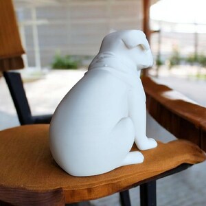 K&ouml;nnte beinhalten: Eine wei&szlig;e Keramik-Bulldoggen-Figur, die auf einem braunen Kissen sitzt. Der Hund ist vom Betrachter abgewandt. Die Figur hat eine glatte, matte Oberfl&auml;che. Der Hund sitzt auf einem Holzstuhl.