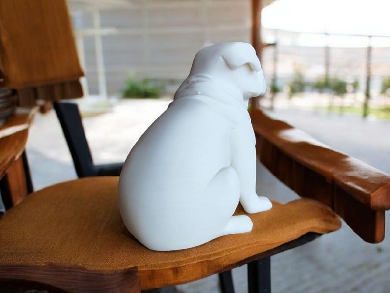 K&ouml;nnte beinhalten: Eine wei&szlig;e Keramik-Bulldoggen-Figur, die auf einem braunen Kissen sitzt. Der Hund ist vom Betrachter abgewandt. Die Figur hat eine glatte, matte Oberfl&auml;che. Der Hund sitzt auf einem Holzstuhl.