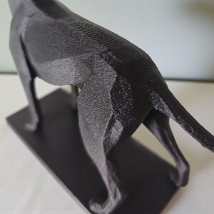 May include: A black, 3D-printed sculpture of a dog, possibly a greyhound, on a rectangular base. The sculpture has a textured surface, with visible layer lines from the printing process. The dog's tail is extended.