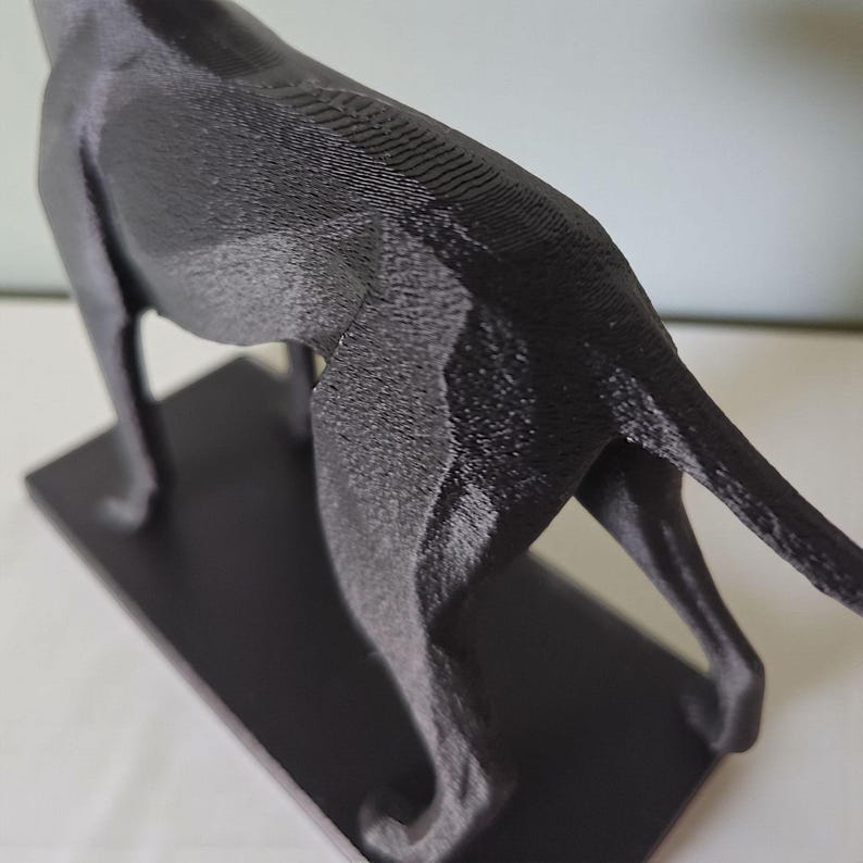 May include: A black, 3D-printed sculpture of a dog, possibly a greyhound, on a rectangular base. The sculpture has a textured surface, with visible layer lines from the printing process. The dog's tail is extended.