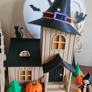May include: A Halloween-themed diorama featuring a witch's hat atop a house with orange windows and a black door. Miniature pumpkins and green trees are in front. A full moon and bats are in the background. A small figure in a black cloak holds a jack-o'-lantern.