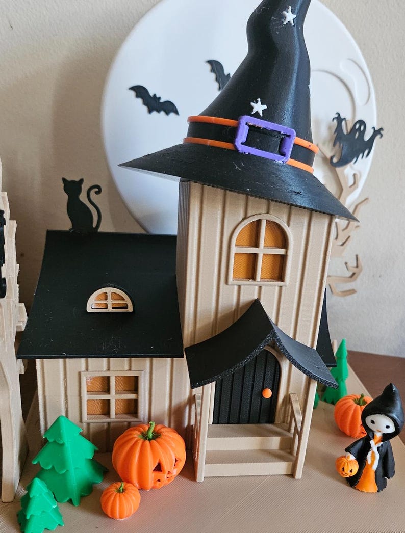 May include: A Halloween-themed diorama featuring a witch's hat atop a house with orange windows and a black door. Miniature pumpkins and green trees are in front. A full moon and bats are in the background. A small figure in a black cloak holds a jack-o'-lantern.