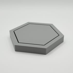 May include: A gray, hexagonal tray with a recessed center. The tray has a geometric design and is made of a solid material. The edges are defined by a thin black line. The tray is suitable for holding small items.