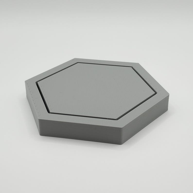 May include: A gray, hexagonal tray with a recessed center. The tray has a geometric design and is made of a solid material. The edges are defined by a thin black line. The tray is suitable for holding small items.
